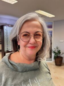 Uśmiechnięta kobieta w średnim wieku z siwymi włosami, nosząca okrągłe okulary w salonie optycznym.